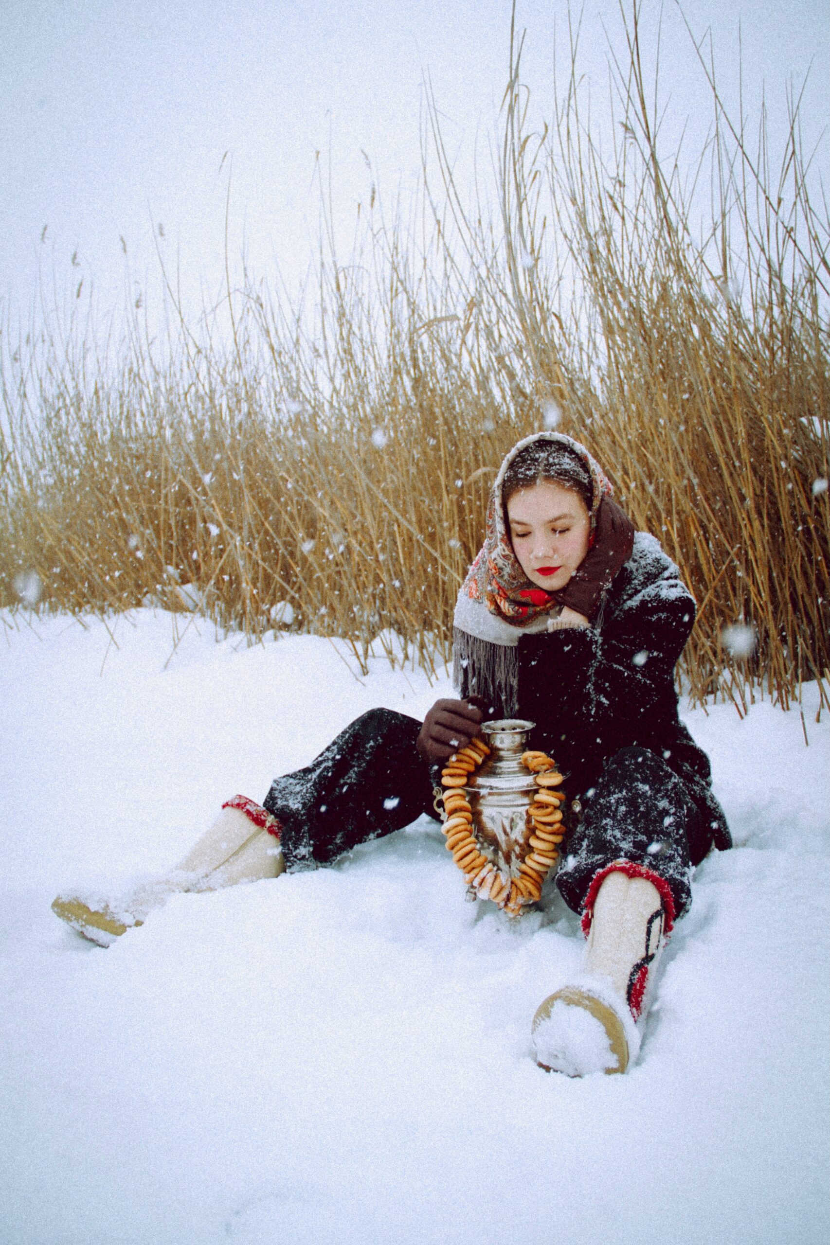 Women in a black jacket, black snow, pants and white boots, and a scarf, sitting in the snow holding an urn